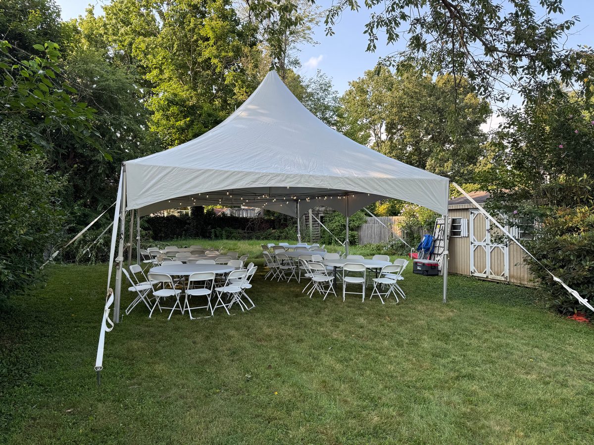 High peak tent with string lights and round tables set up in a leafy Long Island backyard