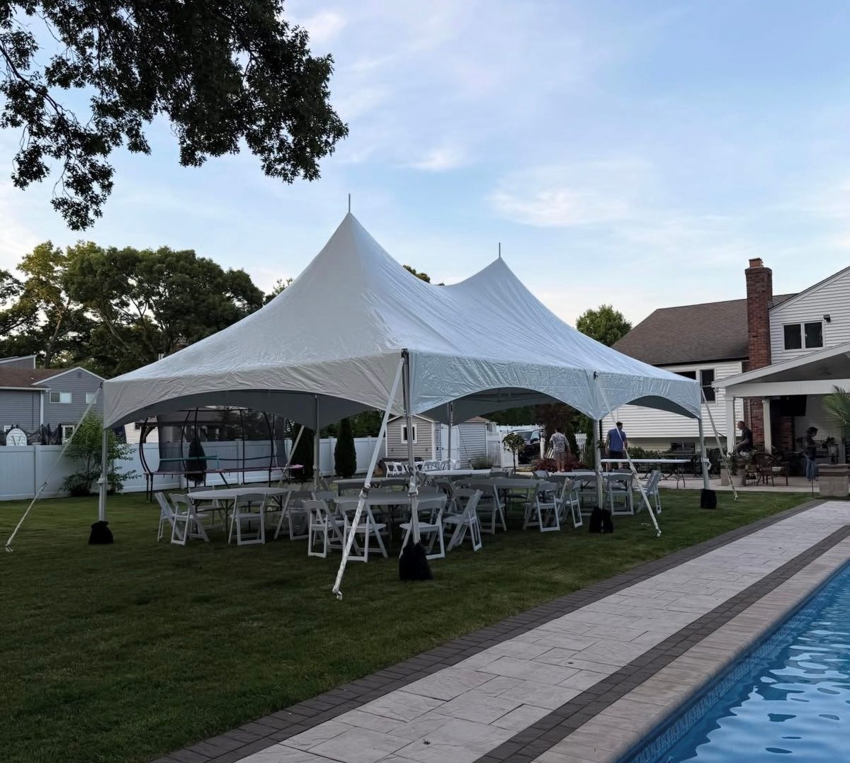 Two-peak party tent with round tables and white chairs next to an in-ground pool and house